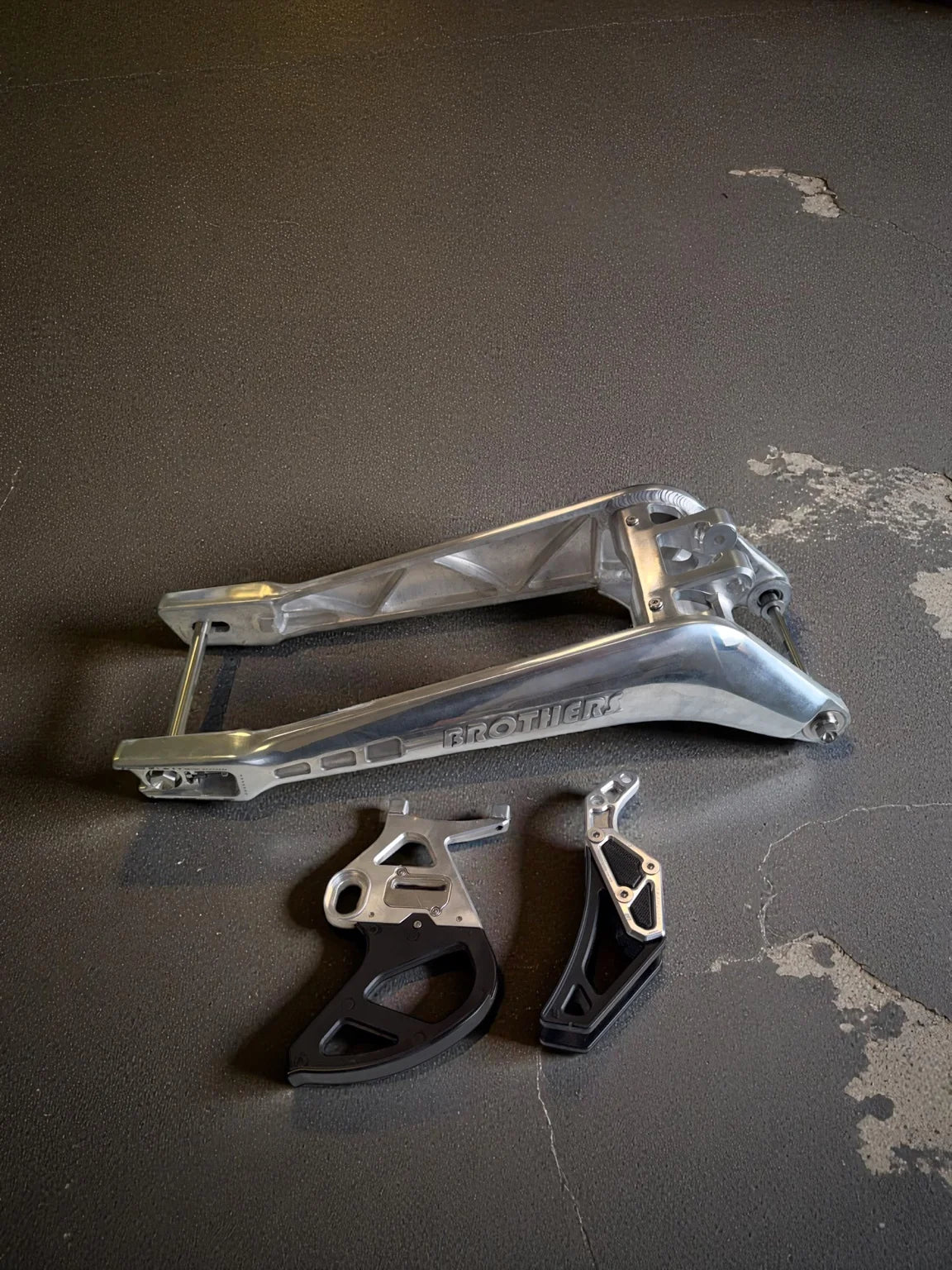 IZI Brothers aluminum swingarm, disc guard, and chain tensioner for Sur-Ron Light Bee displayed on workshop floor